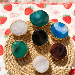 Puede incluir: Un conjunto de siete tiradores de armario de piedras preciosas en varios colores, incluyendo blanco, verde, azul, negro y marrón. Cada tirador tiene un borde dorado. Los tiradores están dispuestos sobre una superficie circular tejida.