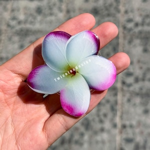 May include: A close-up of a white and purple plumeria flower. The flower has five petals with white centers and edges that fade into a vibrant purple. The flower is held in a hand. The text "TwiceteaDesign" is visible on the flower.