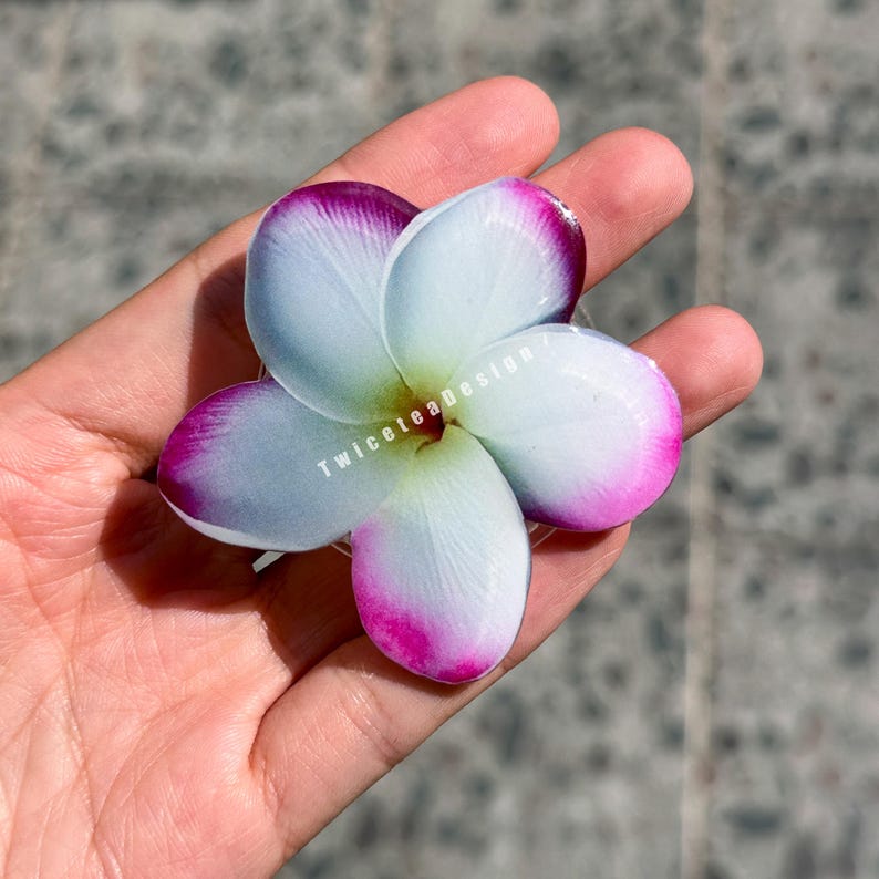 May include: A close-up of a white and purple plumeria flower. The flower has five petals with white centers and edges that fade into a vibrant purple. The flower is held in a hand. The text "TwiceteaDesign" is visible on the flower.