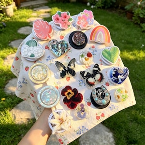 May include: A collection of various circular phone grips featuring diverse designs including floral patterns, butterflies, a rainbow, a heart, a horseshoe, and ocean waves, all arranged on a white fabric cloth with a small strawberry and flower print.