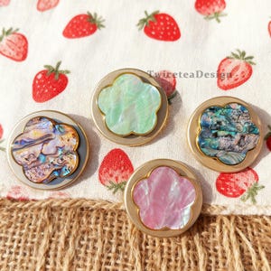 May include: Four decorative flower-shaped buttons with iridescent centers in shades of green, pink, and blue, set in gold-colored metal frames. The buttons are arranged on a strawberry-patterned fabric.