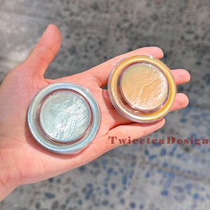Może przedstawiać: Dwa okrągłe, opalizujące kompakty z perłowym wykończeniem trzymane są w dłoni. Jeden jest w kolorze srebrnym, drugi w kolorze złotym. Tekst "TwiceteaDesign" jest widoczny na zdjęciu.