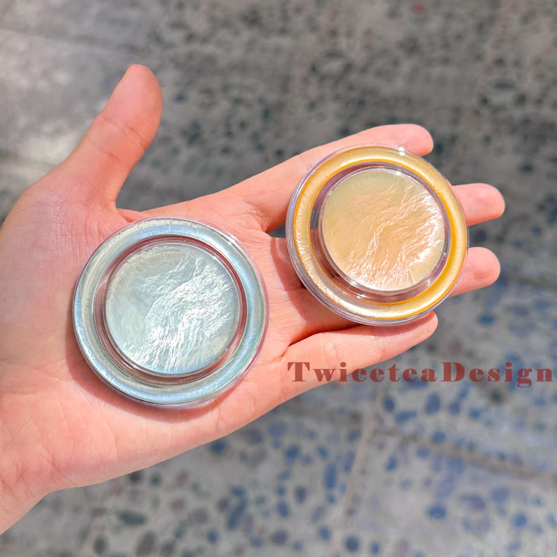 Może przedstawiać: Dwa okrągłe, opalizujące kompakty z perłowym wykończeniem trzymane są w dłoni. Jeden jest w kolorze srebrnym, drugi w kolorze złotym. Tekst "TwiceteaDesign" jest widoczny na zdjęciu.