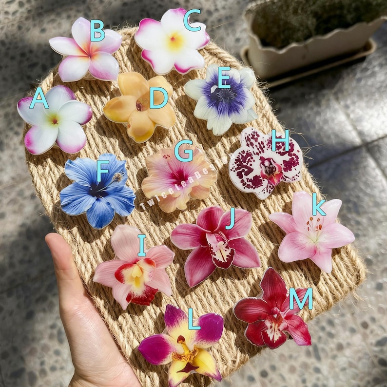 May include: A collection of colorful, flower-shaped accessories, including orchids and hibiscus, arranged on a woven surface. The flowers display a variety of colors, such as pink, white, yellow, and blue. The image is well-lit, showcasing the details of each floral design.