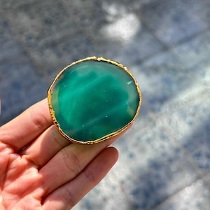 Può includere: Una pietra di agata verde lucida e rotonda con un bordo dorato. La pietra è tenuta in mano, mostrando le sue variazioni di colore naturali e la sua superficie liscia. Il bordo dorato aggiunge un tocco di eleganza alla pietra naturale.