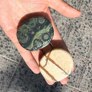 Könnte beinhalten: Zwei polierte Steine werden in einer Hand gehalten. Einer ist dunkelgrün mit helleren grünen und beigen Mustern, der andere ist beige mit braunen Adern. Der Text "twiceagestudio" ist auf dem grünen Stein sichtbar.