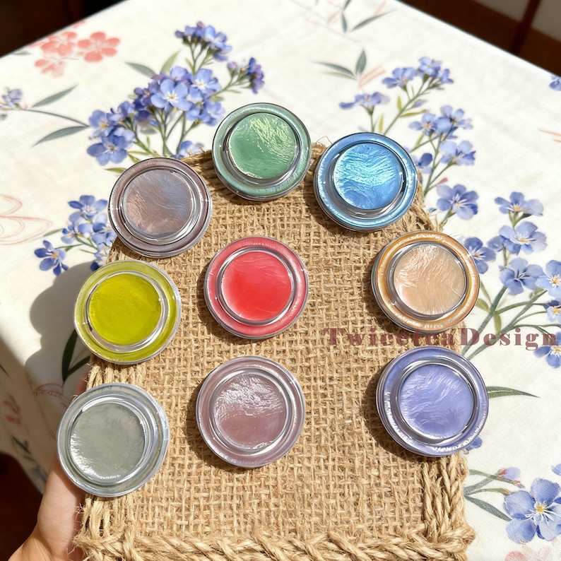 Może przedstawiać: Kolekcja dziewięciu okrągłych, iryzujących kompakt&oacute;w w r&oacute;żnych kolorach, w tym zielonym, niebieskim, r&oacute;żowym i ż&oacute;łtym. Każdy kompakt ma srebrną obw&oacute;dkę i połyskujące, perłowe wykończenie. Kompakty są ułożone na jutowej powierzchni.