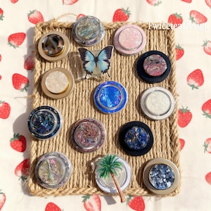May include: A selection of decorative phone grips featuring diverse designs, such as iridescent, marble, and butterfly patterns. The grips are displayed on a woven jute placemat, set against a strawberry-patterned backdrop.