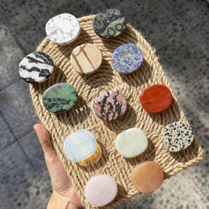 Op de afbeelding: Een collectie van twaalf ronde telefoonhouders met diverse steenachtige patronen en effen kleuren, waaronder wit marmer, zwart-witte strepen, groen, roze, blauw, rood en doorschijnende afwerkingen, gerangschikt op een displaybord van gevlochten touw.