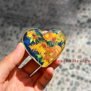 May include: Heart-shaped decorative item with a gold-colored border. The interior features a vibrant floral design of sunflowers in shades of yellow, orange, and green against a dark blue background. The text "TwiceteaDesign" is visible.