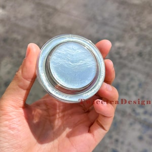 Może przedstawiać: Przezroczysty, okrągły pojemnik z perłową, jasnoniebieską substancją w środku. Pojemnik ma srebrną obw&oacute;dkę i jest trzymany w dłoni. Produkt jest prawdopodobnie kosmetykiem lub produktem do pielęgnacji sk&oacute;ry.