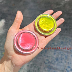 Może przedstawiać: Dwa okrągłe, przezroczyste pojemniki z kolorową zawartością trzymane w dłoni. Jeden pojemnik ma r&oacute;żowe wnętrze, a drugi ż&oacute;łte. Pojemniki mają przezroczystą, opalizującą obw&oacute;dkę. Widoczny jest tekst "TwiceteaDesign".
