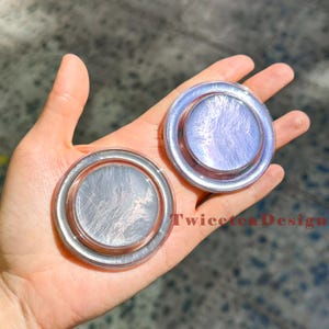 Może przedstawiać: Dwa iryzujące, okrągłe przedmioty dekoracyjne z perłowym wykończeniem. Obiekty mają srebrne i r&oacute;żowo-złote obramowanie. Tekst "Twicetea Design" jest widoczny na zdjęciu.