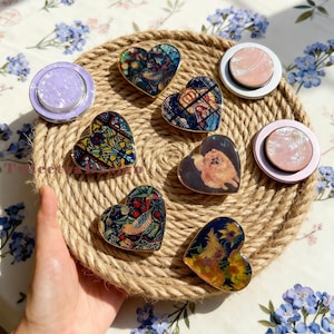 May include: A collection of heart-shaped phone grips and round phone grips with various designs. The heart-shaped grips feature colorful patterns and a dog image. The round grips have a pearlescent finish. The items are arranged on a woven mat.