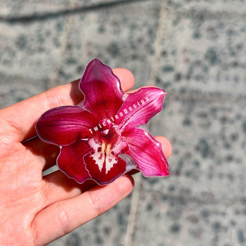 May include: A vibrant, resin-cast orchid flower in shades of deep red and pink, with a white and yellow center. The flower is held in a hand, and the text "Twiceteadesign" is visible on the petals.