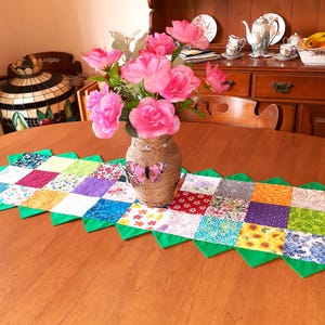 May include: A patchwork table runner with a green scalloped edge. A vase with a pink butterfly design holds a bouquet of pink flowers.