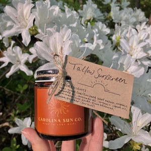 May include: A brown glass jar with a gold label that reads "CAROLINA SUN CO. kind to your body". The jar contains a brown substance and is labeled "Tallow Sunscreen" with a list of ingredients: 100% grass fed/finished beef tallow, organic coconut oil, avocado oil, organic jojoba oil, non-nano zinc oxide, pure beeswax, Cocoa powder (for tint).