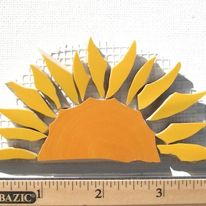May include: A mosaic sun design with a bright orange center and yellow rays. The sun is made of small, irregular pieces, creating a textured effect. The design is approximately 3 inches wide, placed on a white surface.