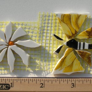 May include: A mosaic art piece featuring a white daisy with an orange center and a yellow and brown butterfly. The artwork is made from broken pieces of ceramic tile. The daisy is approximately 1.5 inches in diameter, and the butterfly is about 2 inches long.