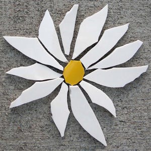 May include: A mosaic flower design composed of individual white ceramic shards arranged as petals radiating from a single, central, hexagonal yellow ceramic piece. The pieces are shaped with jagged, irregular edges to form a daisy-like floral pattern.