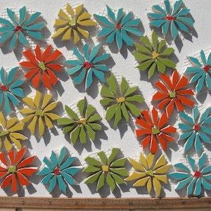 May include: A collection of twenty-one flower-shaped mosaic tiles in shades of turquoise, orange, lime green, and yellow, each featuring a small contrasting center piece, arranged on a white mesh backing above a wooden ruler measuring 10 inches.