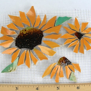 May include: Three mosaic sunflowers with orange petals and dark brown centers. The mosaic pieces are arranged on a white grid. The sunflowers vary in size, with one large and two smaller ones.
