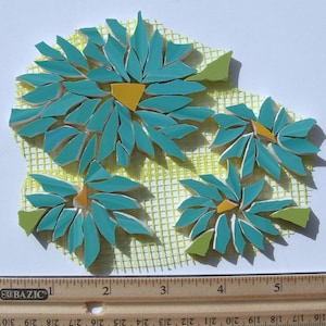 May include: A mosaic art project featuring four flower shapes made of teal ceramic pieces with yellow centers, arranged on a yellow mesh backing. A wooden ruler is positioned at the bottom, showing the project width is approximately 5.5 inches.