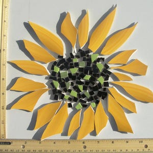 May include: A sunflower mosaic art piece composed of yellow, black, and light green glass tiles. A wooden ruler is positioned along the left and bottom edges, showing the piece measures approximately 11 inches in width and 11 inches in height.