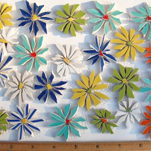 May include: A collection of mosaic flower tiles in various colors, including yellow, blue, green, white, and orange. Each tile has a central accent and radiating petals, creating a decorative floral design. The tiles are arranged on a white surface.