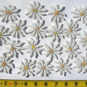 May include: A collection of white mosaic daisy flower tiles with yellow and orange centers, arranged on a white surface. The mosaic tiles are on a mesh backing. A yellow measuring tape is at the bottom of the image.