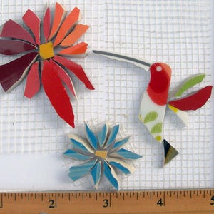 May include: Mosaic art pieces of a red, orange, and yellow flower, a blue and yellow flower, and a hummingbird with red, white, green, and yellow details. The pieces are on a white grid, with a wooden ruler for scale.