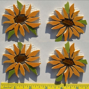 May include: Four mosaic sunflower designs. Each sunflower has orange petals, a brown center, and green leaves. The sunflowers are set on a white background. A yellow ruler with black markings is at the bottom.