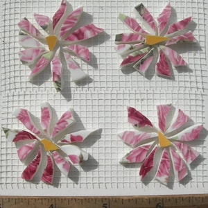 May include: Four mosaic flower tiles with pink floral patterns and yellow centers are arranged on a white grid. The petals are made of broken ceramic pieces. A ruler is at the bottom.