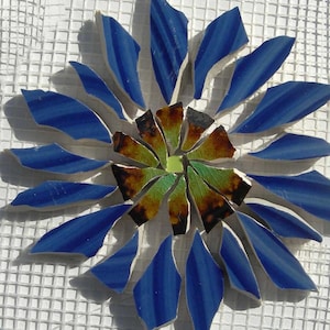 May include: A mosaic flower design with blue and green glass pieces. The petals are a vibrant blue with darker blue stripes, radiating from a center of green and brown pieces. The mosaic is set on a white grid background.