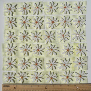 May include: Arrangement of white daisy-shaped mosaic tiles with orange centers, set against a light yellow grid. A wooden ruler with inch measurements is at the bottom. The tiles are arranged in rows.