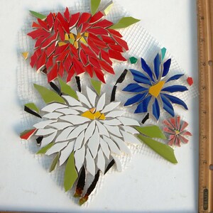 May include: A mosaic art piece featuring three floral designs in red, white, and blue glass tiles with green leaf accents, arranged on a white mesh backing. A wooden ruler is visible along the right and bottom edges, showing a width of 9 inches.