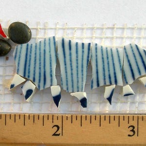 May include: A mosaic art piece featuring blue and white striped ceramic shards arranged on a mesh backing. Two dark green stones and a red and yellow triangular piece are also included. A wooden ruler with inch markings is in the foreground.