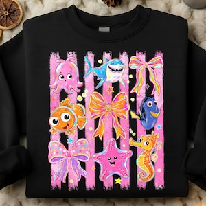 May include: Black sweatshirt featuring a colorful graphic of cartoon sea creatures and bows. The design includes an octopus, shark, clownfish, bow, starfish, seahorse, and a blue fish. The background has pink vertical stripes.