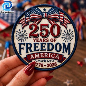 Może przedstawiać: Okrągła haftowana naszywka z czerwoną, białą i niebieską kokardą w stylu amerykańskiej flagi. Napis 250 YEARS OF FREEDOM AMERICA 1776-2026 wykonano czerwoną i niebieską nicią. Wzór zawiera motywy fajerwerków na kremowym tle.
