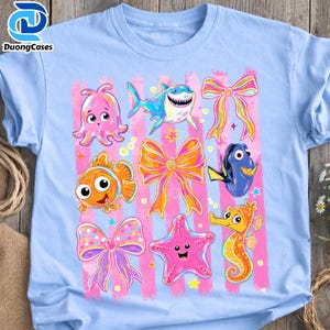 May include: Light blue t-shirt featuring a colorful graphic of cartoon sea creatures, bows, and stars. The design includes an octopus, shark, clownfish, bow, Dory, starfish, and seahorse. The shirt has pink vertical stripes.