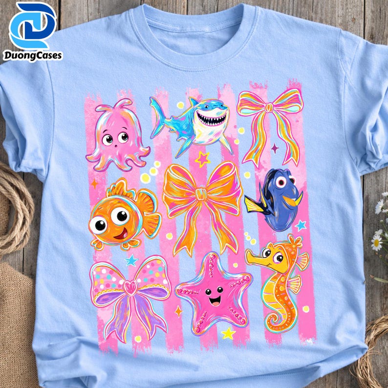 May include: Light blue t-shirt featuring a colorful graphic of cartoon sea creatures, bows, and stars. The design includes an octopus, shark, clownfish, bow, Dory, starfish, and seahorse. The shirt has pink vertical stripes.