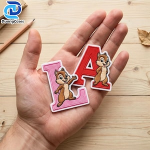May include: Two embroidered fabric patches shaped like the letters L and A, each featuring a brown and tan chipmunk character. The L patch is pink with a white border, and the A patch is red with a white border, both with detailed stitching.