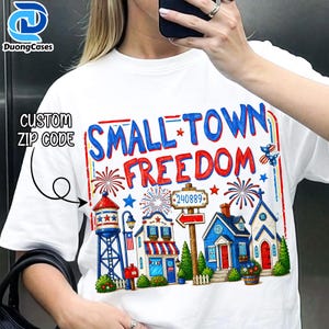 以下が含まれることがあります： 白のTシャツ。赤と青で「SMALL TOWN FREEDOM」の文字が描かれたカラフルなグラフィックデザイン。デザインには、給水塔、家、花火、郵便番号240889の看板のイラストが含まれています。