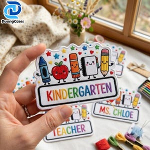 May include: A white embroidered patch featuring a colourful school theme with a blue crayon, red apple, orange marker, white notebook, and yellow pencil. The word KINDERGARTEN is stitched in rainbow letters on a white rectangular background.