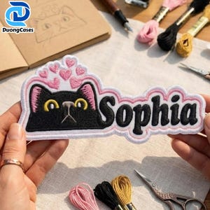 May include: An embroidered patch featuring a black cat with yellow eyes and pink inner ears, surrounded by small pink hearts above its head. The name Sophia is stitched in black letters to the right of the cat, all outlined in pink thread.
