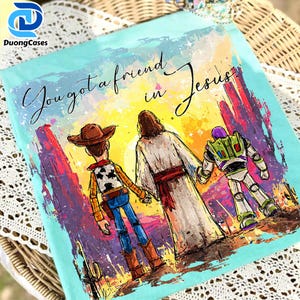 Può includere: Maglietta azzurra con un'illustrazione colorata di Woody, Gesù e Buzz Lightyear che si tengono per mano. Il testo "You got a friend in Jesus" è scritto sopra. L'opera d'arte è in stile acquerello, con uno sfondo di tramonto.