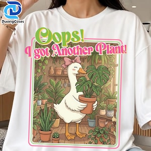 Pode incluir: Camiseta branca com um ganso de desenho animado com um laço rosa segurando uma planta em vaso. A imagem tem o texto "Oops! I got Another Plant!" em verde e rosa. O fundo mostra uma loja de plantas com várias plantas em vasos.