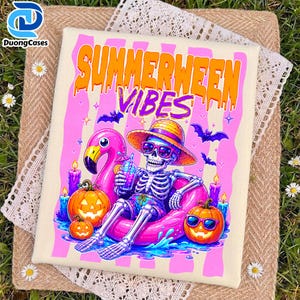 Peut inclure: T-shirt crème avec un graphisme coloré. Le motif représente un squelette portant des lunettes de soleil et un chapeau, se relaxant sur un flotteur flamant rose. Le texte "SUMMERWEEN VIBES" est affiché au-dessus du squelette, avec des citrouilles et des chauves-souris.