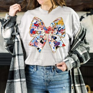 K&ouml;nnte beinhalten: Ein wei&szlig;es Langarmshirt mit einer rot-wei&szlig;-blauen Schleife mit Mickey Mouse und anderen Disney-Charakteren. Die Schleife ist mit Sternen und Streifen verziert.