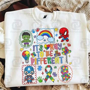 May include: White sweatshirt with a colorful graphic that reads "It's Okay to Be Different." The design includes a rainbow, puzzle pieces, and cartoon characters like Hulk, Spider-Man, and Captain America. The sweatshirt is a crew neck style.